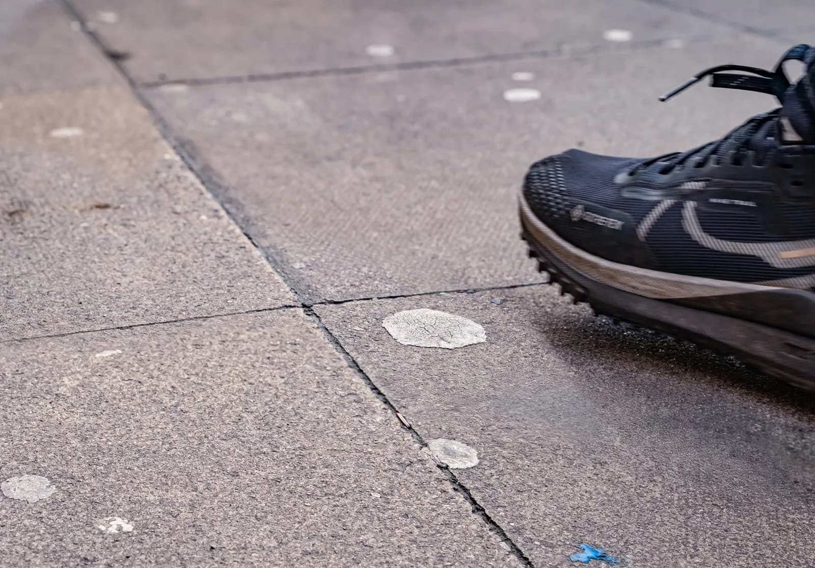 A close-up view of a dark grey running shoe with a rugged sole stepping on a textured concrete pavement littered with flattened, white chewing gum blobs.