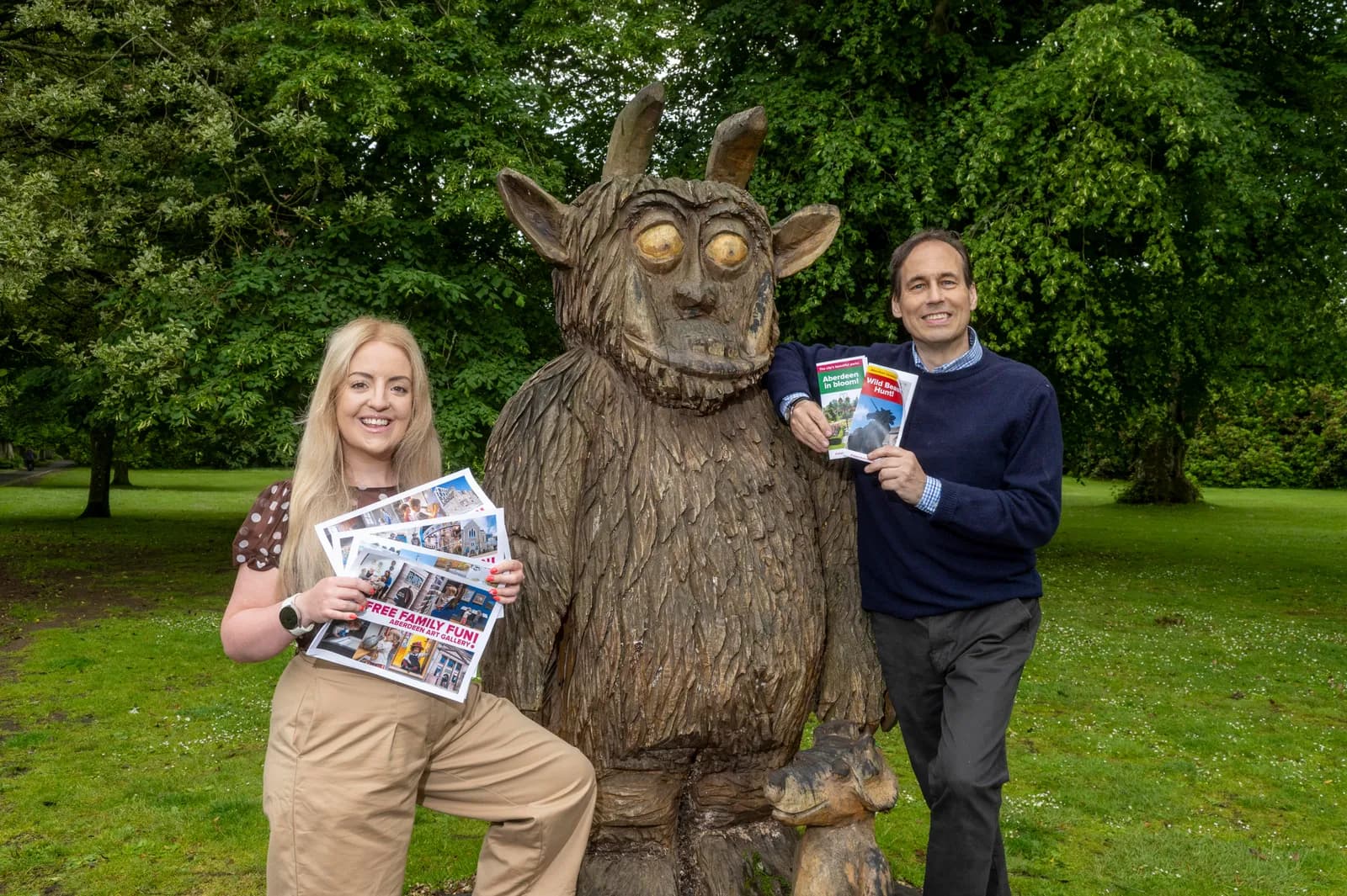 A woman and a man holding magazines, standing next to a large wooden sculpture in a park.