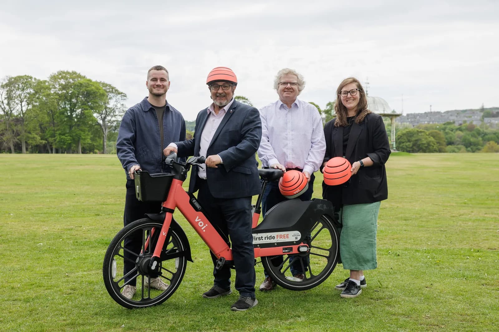 Four people, two men and two women, one of whom is wearing an orange helmet, stand around an orange Voi electric bicycle on a grassy field, holding two smaller orange helmets.