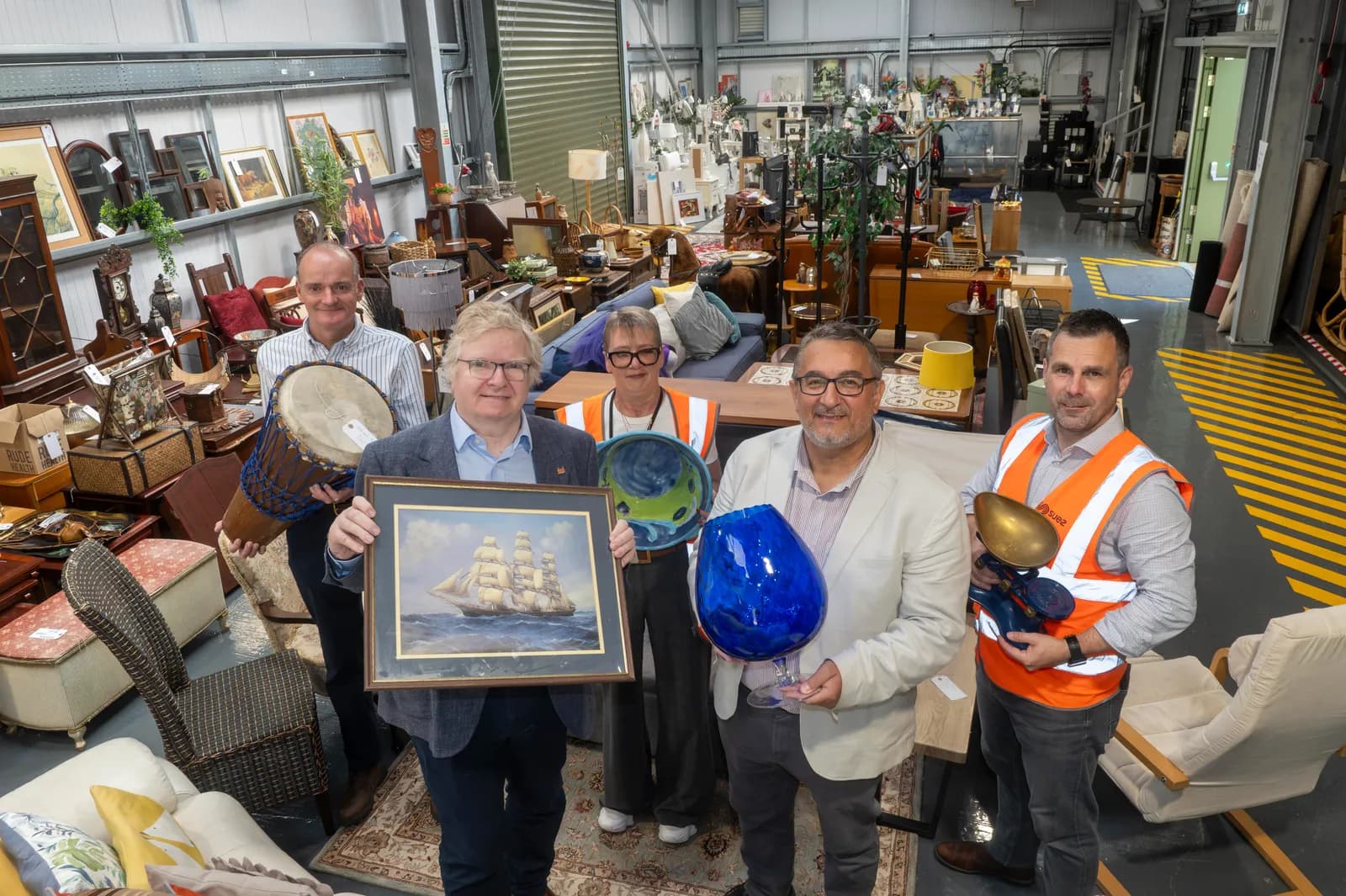Five people in a warehouse filled with various items, including furniture, paintings, and decorative objects, hold up an array of items.