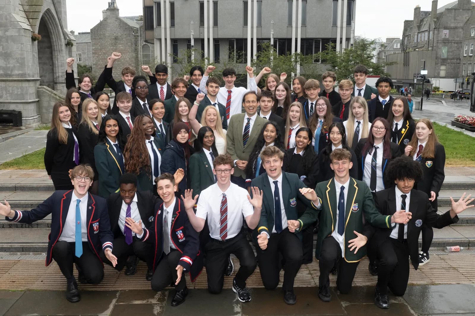 A diverse group of students in school uniforms pose for a photo outdoors, many with arms raised in celebration, with a teacher among them.