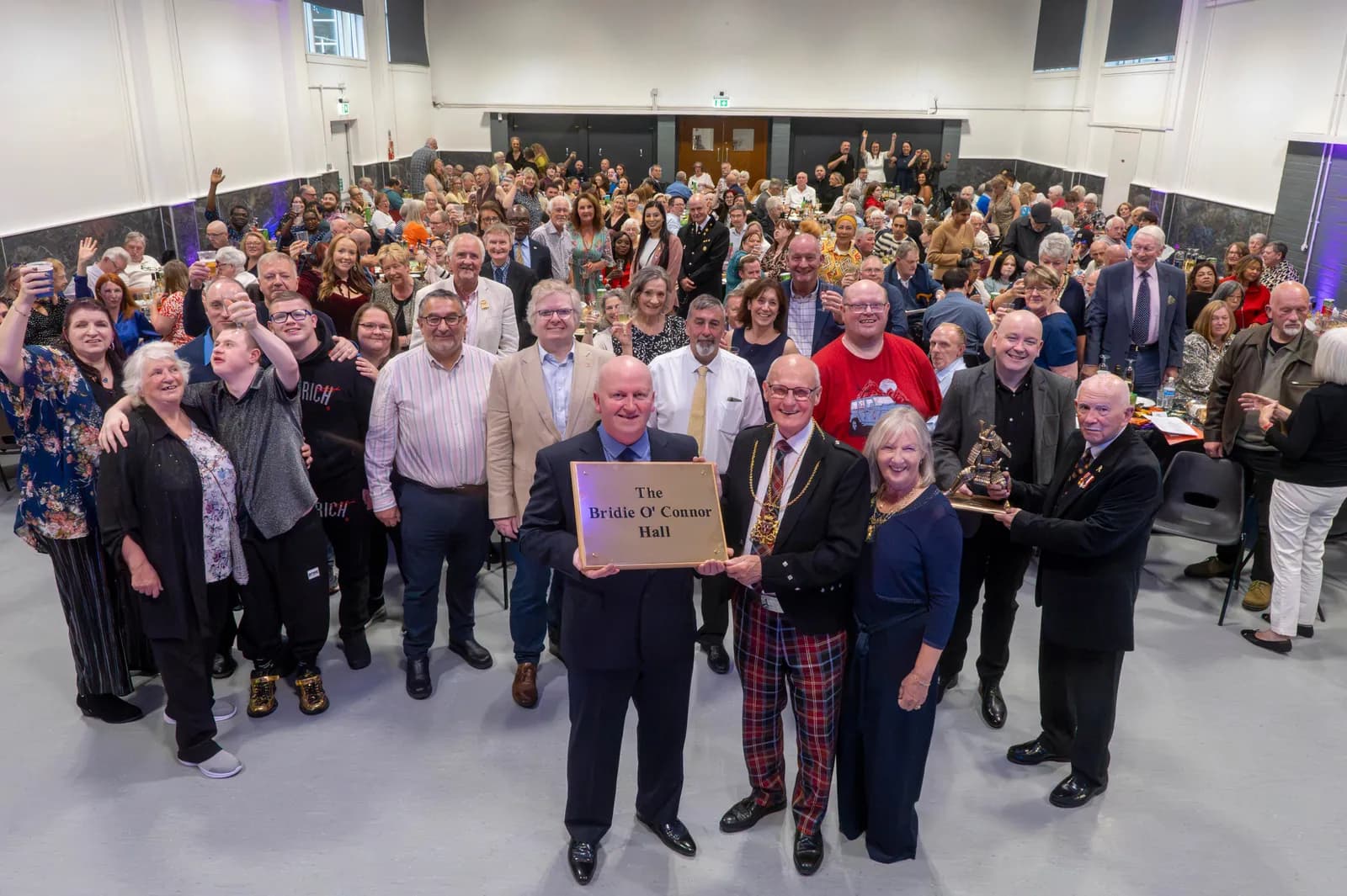 A large group of people gathered in a hall, with a few individuals in the foreground holding awards and a plaque that reads "The Bridie O' Connor Hall."