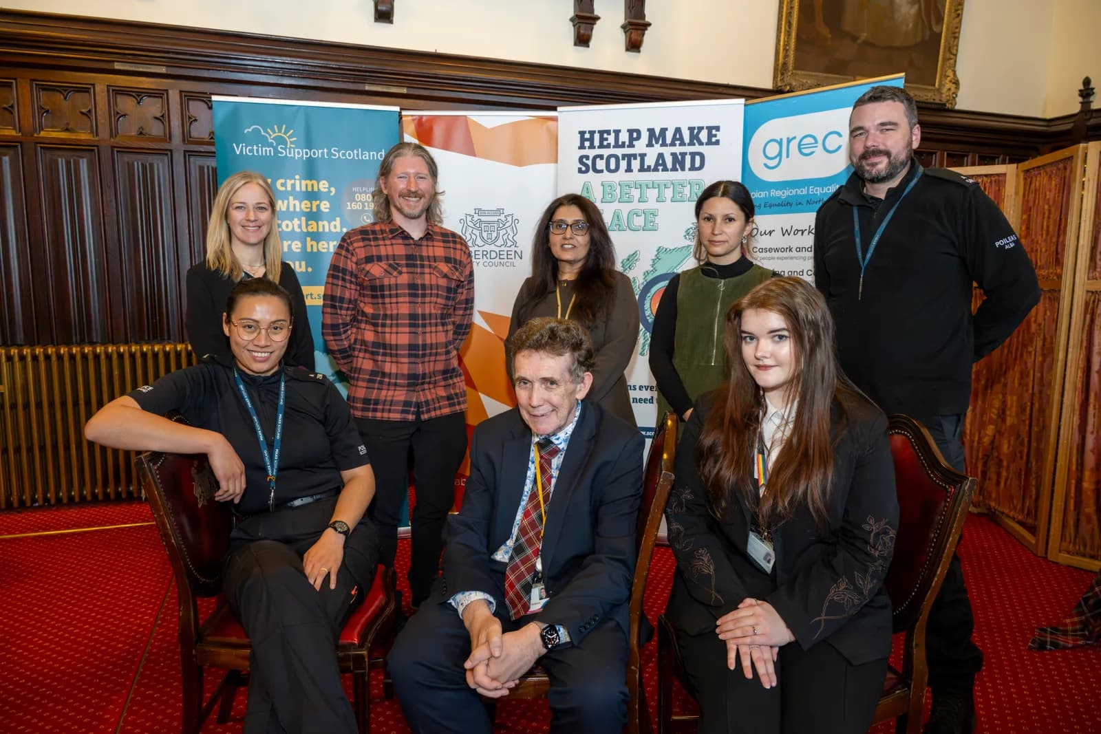 A group of eight individuals, some standing and some sitting, smile in front of banners promoting organizations like Victim Support Scotland and Help Make Scotland a Better Place.