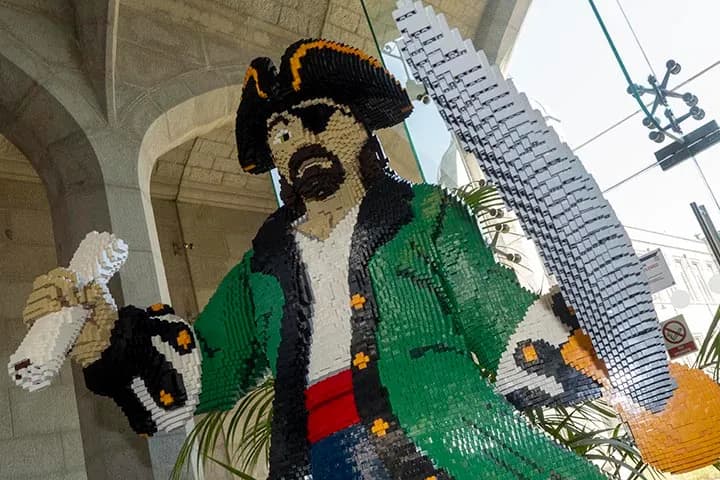 A large, colorful sculpture of a pirate made entirely of Lego bricks, wearing a green coat, black hat, and eye patch, holding a white parrot on one arm and a sword in the other hand, inside a building with stone arches and large windows.