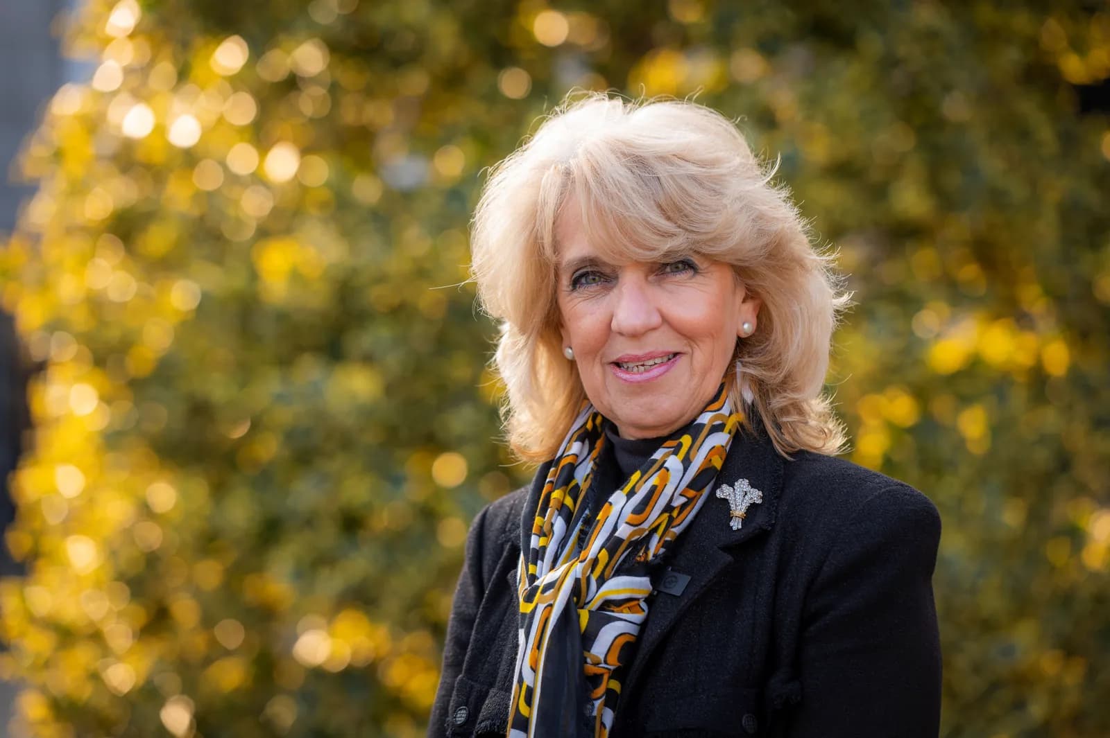 A smiling woman with blonde hair, a dark jacket, and a colorful scarf stands in front of a blurred background of green and yellow foliage.