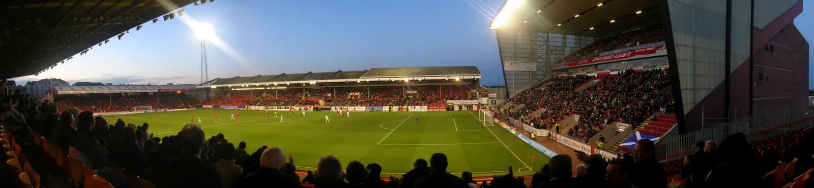 A wide-angle view of a football stadium from the stands, showing a match in progress on a brightly lit green field with a large crowd in the spectator seating.