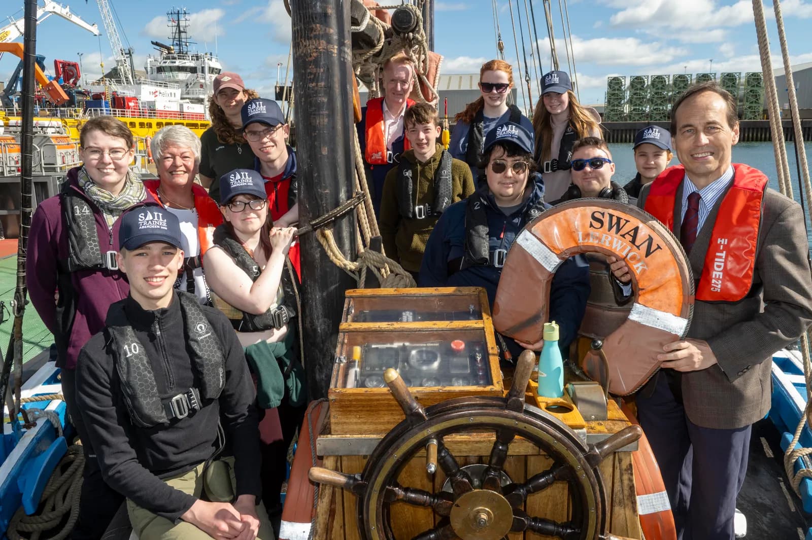 A group of young people, and some adults, pose on the deck of a sailboat, with a man in a business suit holding a life preserver in the foreground.